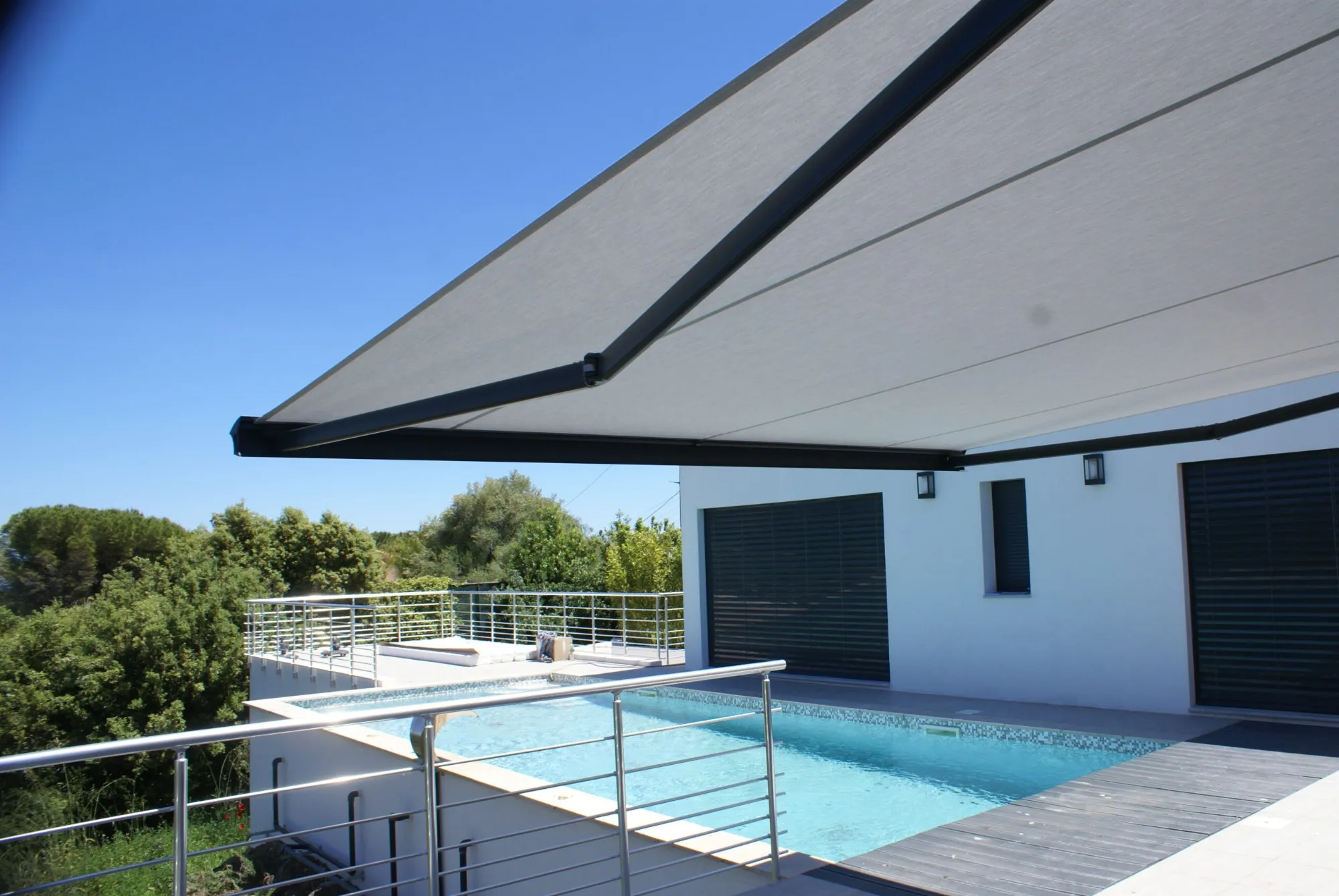 Store banne gris vu de dessous, bras articulés déployés au-dessus d’une terrasse dominant une piscine